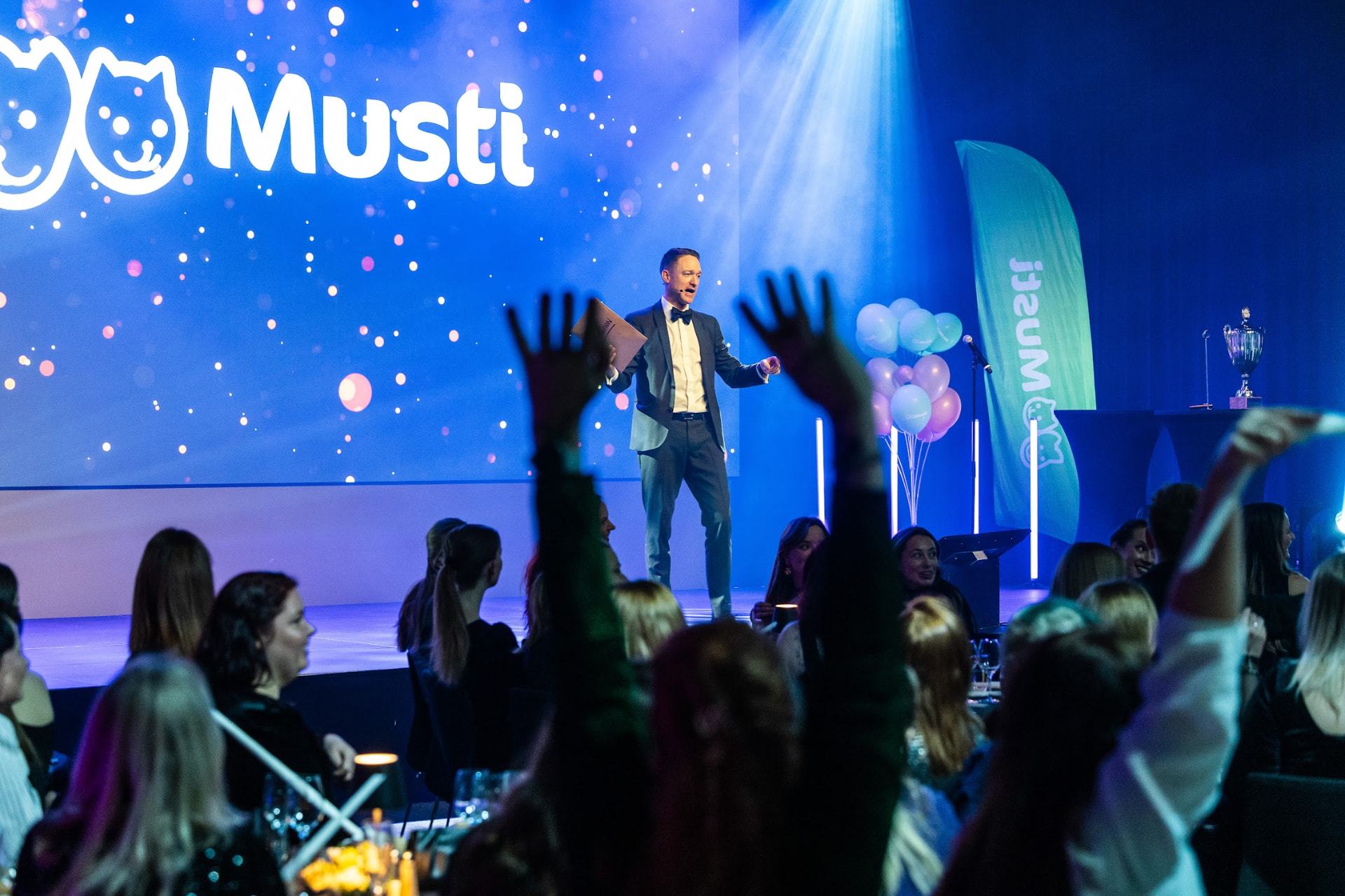 Deltakere rekker opp hendene foran scenen med Musti-logoen