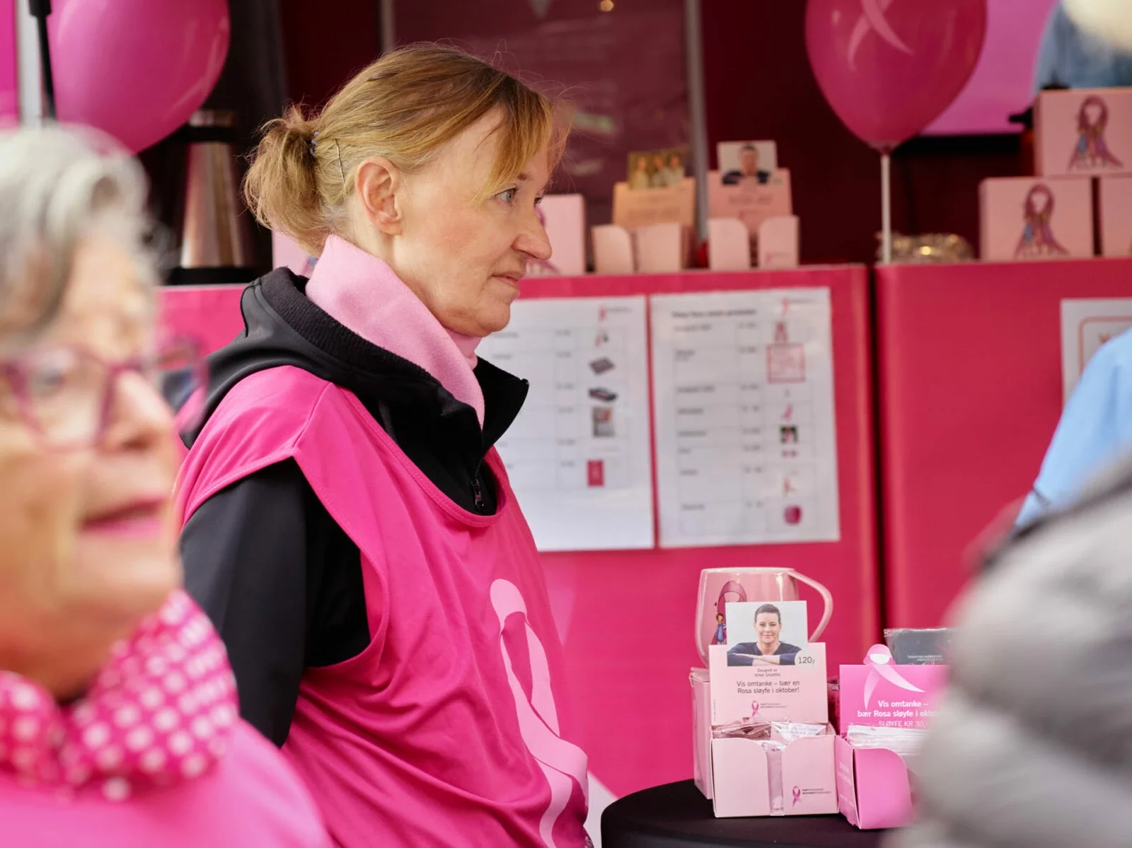 Person i rosa vest ved standen, med besøkende i forgrunnen.