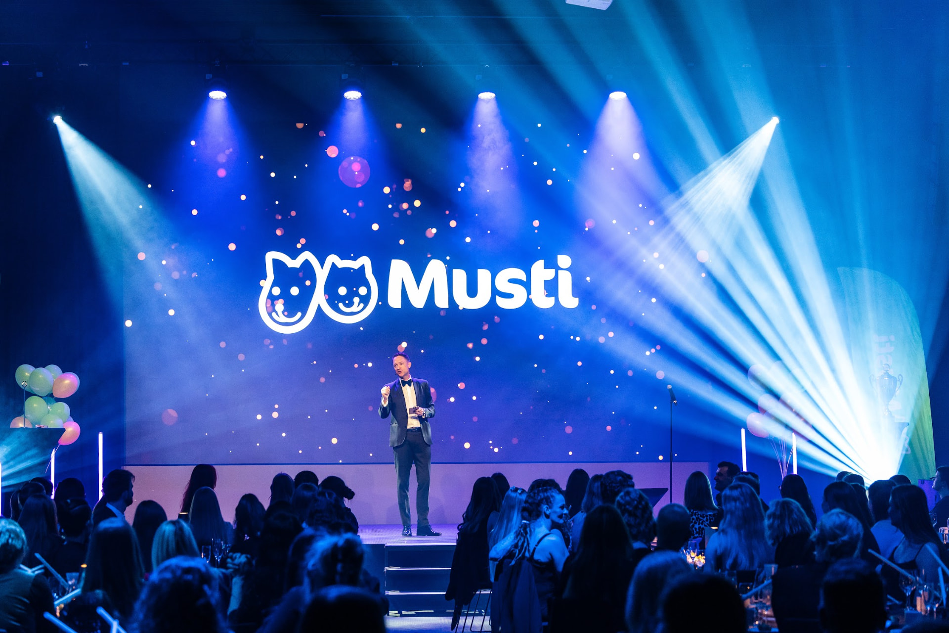 Konferansier på scenen foran storskjerm med Musti-logo og publikum i salen