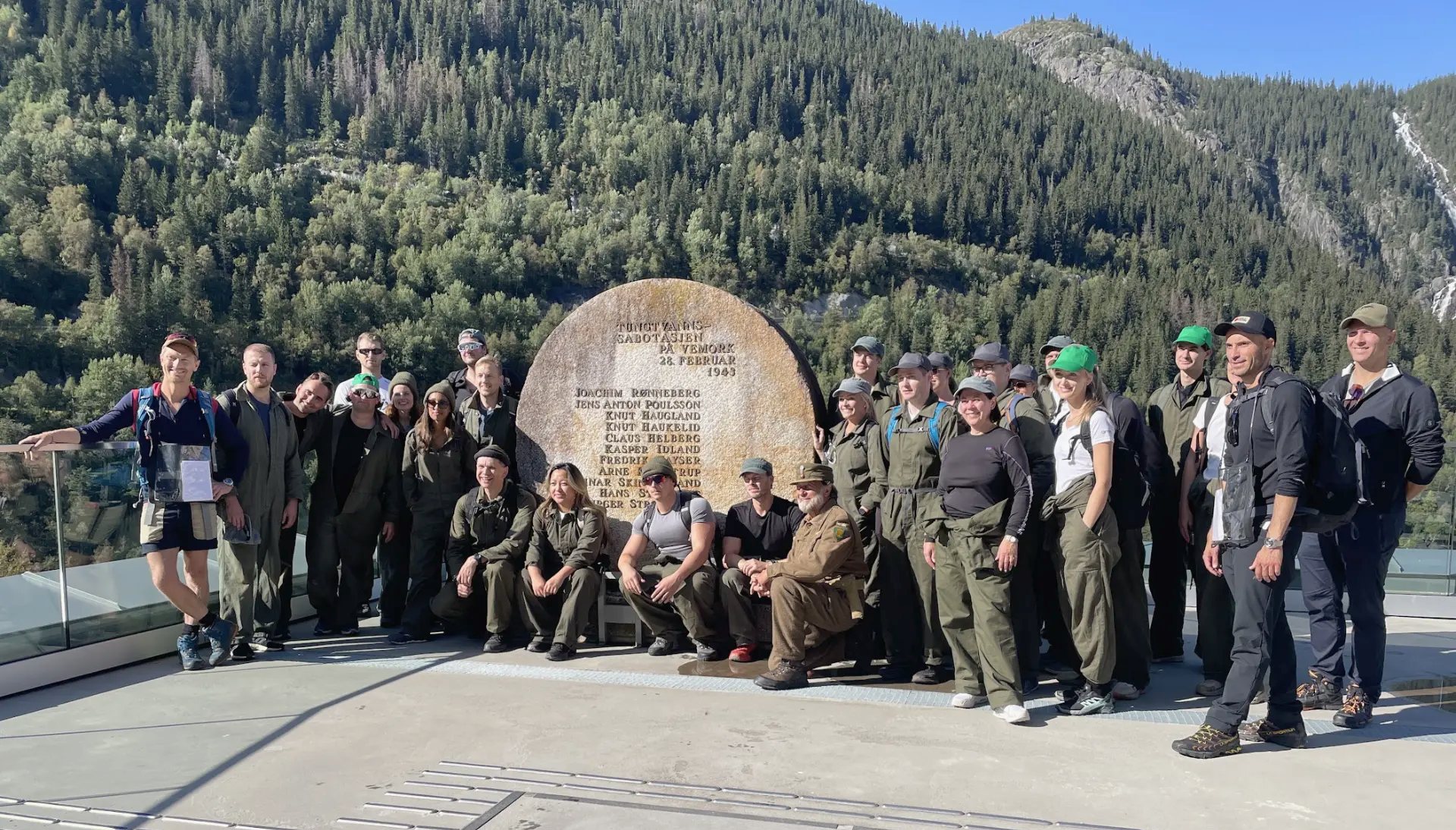 Gruppen samlet ved et minnesmerke i stein med skogkledde fjell bak