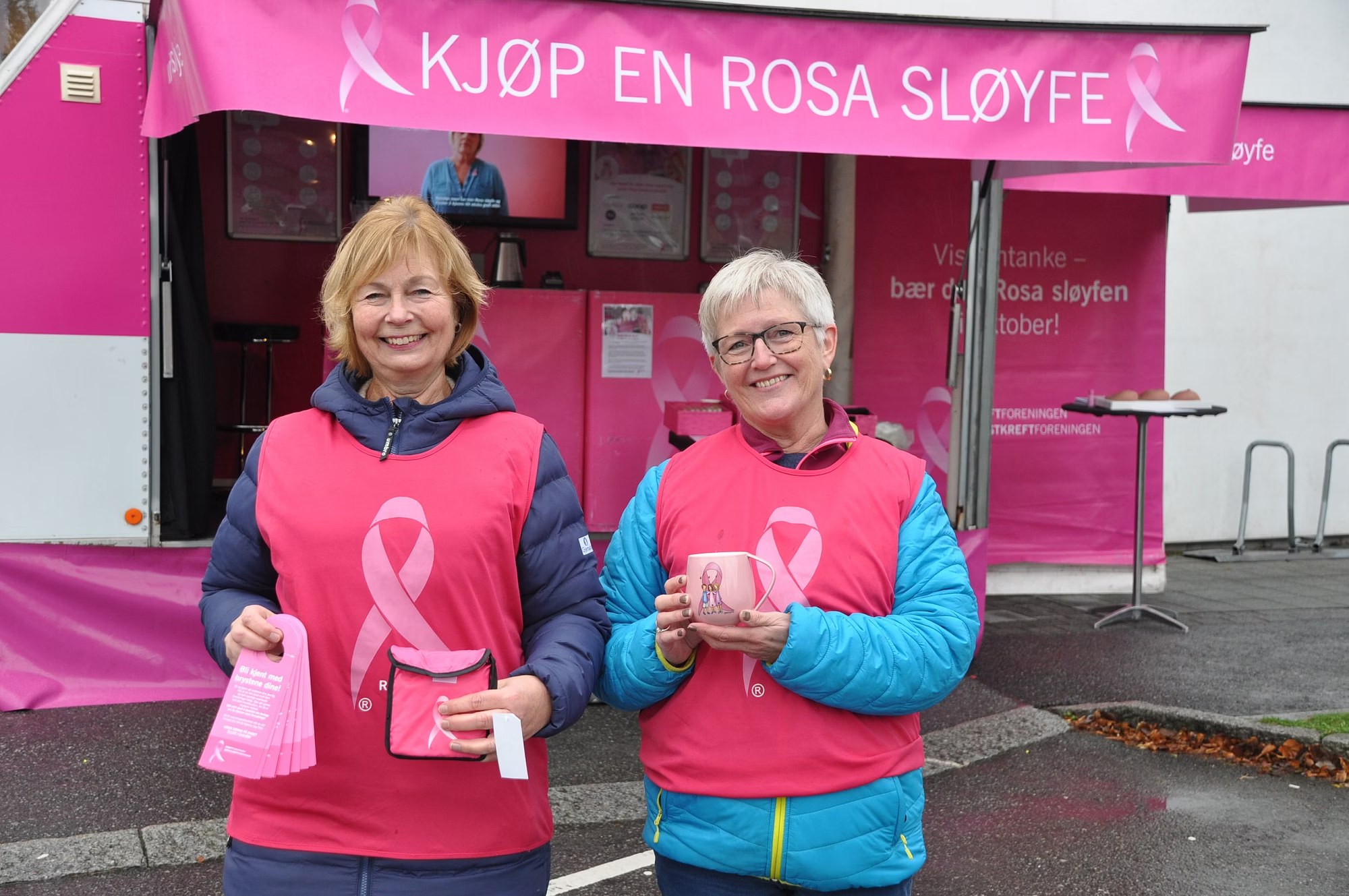 To kvinner i rosa vester med produkter foran «Kjøp en rosa sløyfe»-markisen.