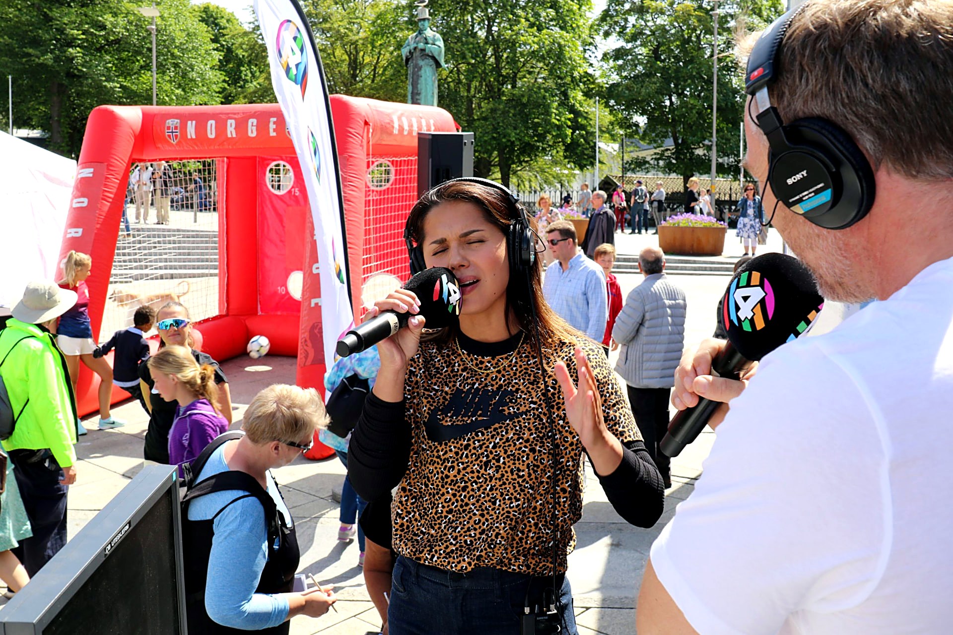 Programleder synger i mikrofon under en direktesending, med publikum og kolleger i bakgrunnen